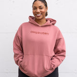 Woman standing facing forward with hands inside her front pocket, wearing a unisex oversize bamboo-organic cotton fleece hoodie in dusty rose, with dropped shoulders and a large kangaroo pocket, featuring the A$$piration Project logo on the front in tone on tone dark ruby pink.