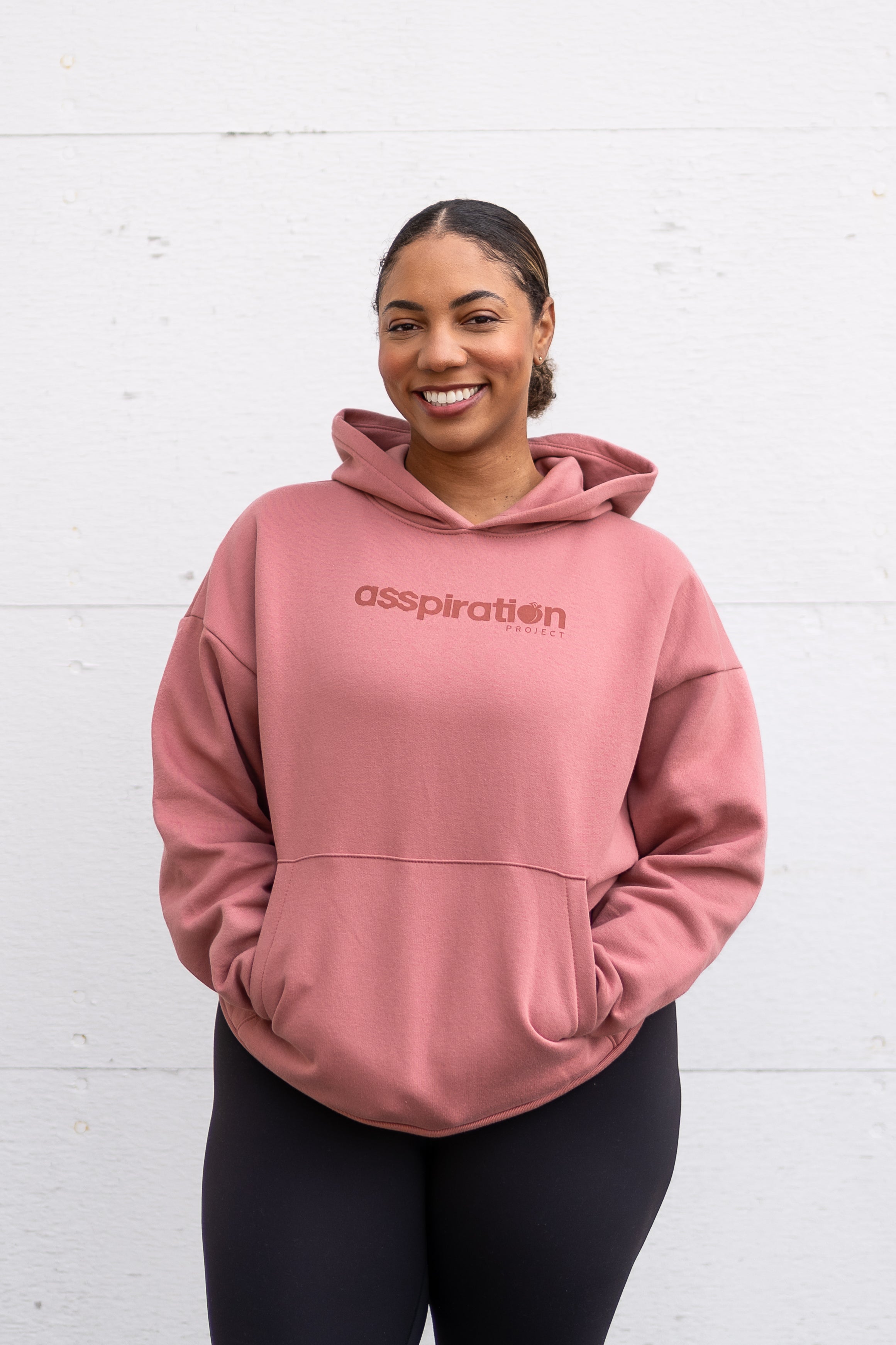 Woman standing facing forward with hands inside her front pocket, wearing a unisex oversize bamboo-organic cotton fleece hoodie in dusty rose, with dropped shoulders and a large kangaroo pocket, featuring the A$$piration Project logo on the front in tone on tone dark ruby pink.