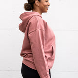Woman standing facing sideways wearing a unisex oversize bamboo-organic cotton fleece hoodie in dusty rose, with dropped shoulders and a large kangaroo pocket, featuring the A$$piration Project logo on the front in tone on tone dark ruby pink.