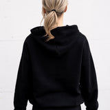 Woman standing facing backwards wearing a unisex oversize bamboo-organic cotton fleece hoodie in black, with dropped shoulders and a large kangaroo pocket.