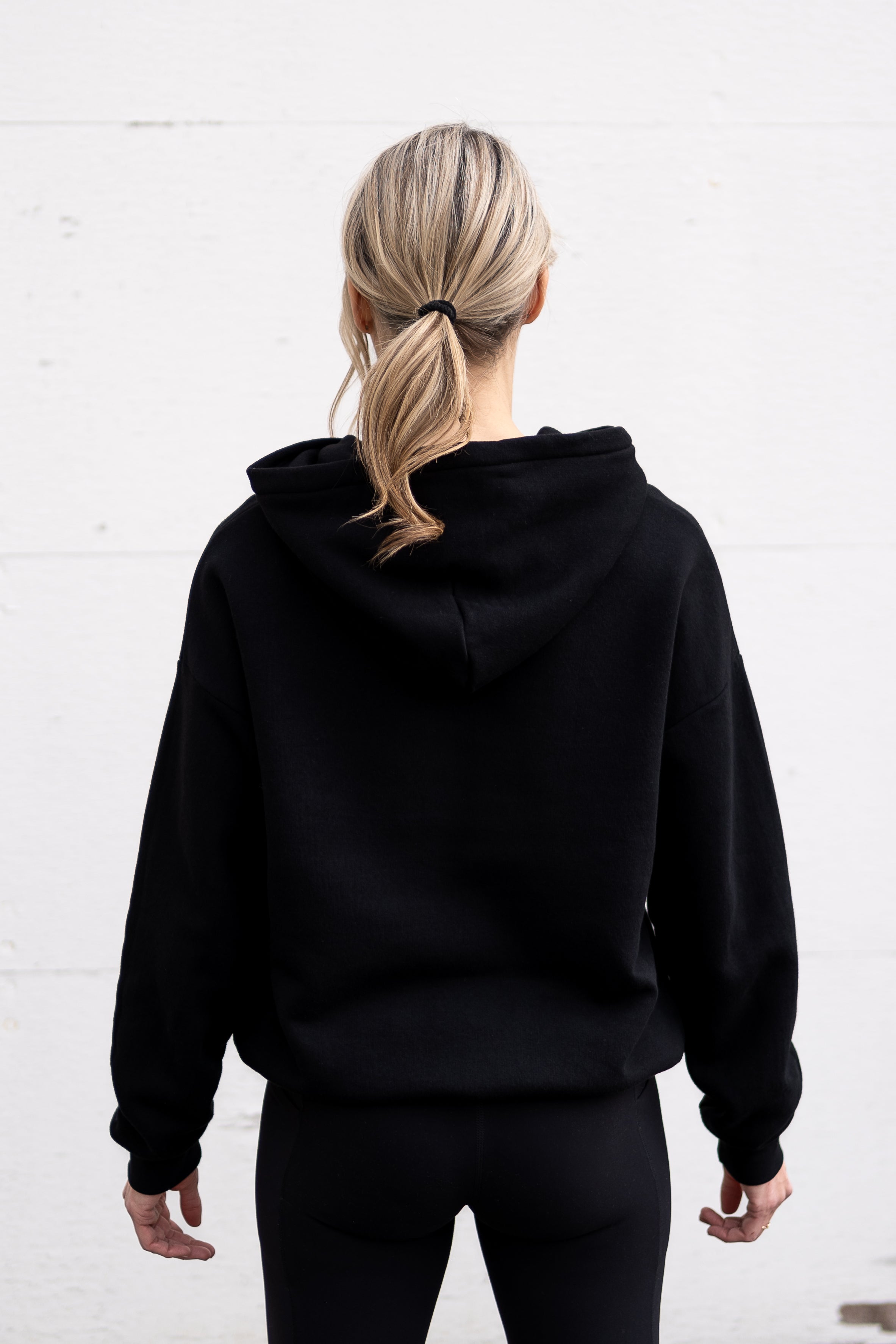 Woman standing facing backwards wearing a unisex oversize bamboo-organic cotton fleece hoodie in black, with dropped shoulders and a large kangaroo pocket.