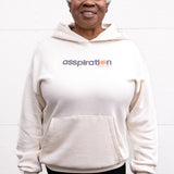Woman standing facing forward with arms at her sides, wearing a unisex oversize bamboo-organic cotton fleece hoodie in cream, with dropped shoulders and a large kangaroo pocket, featuring the A$$piration Project logo on the front in grey, peach and green.
