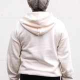 Woman standing facing forward with arms at her sides, wearing a unisex oversize bamboo-organic cotton fleece hoodie in cream, with dropped shoulders and a large kangaroo pocket.