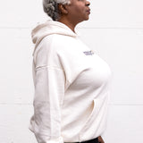 Woman standing facing sideways with arms at her sides, wearing a unisex oversize bamboo-organic cotton fleece hoodie in cream, with dropped shoulders and a large kangaroo pocket, featuring the A$$piration Project logo on the front in grey, peach and green.
