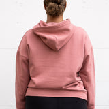 Woman standing facing backwards wearing a unisex oversize bamboo-organic cotton fleece hoodie in dusty rose, with dropped shoulders and a large kangaroo pocket.