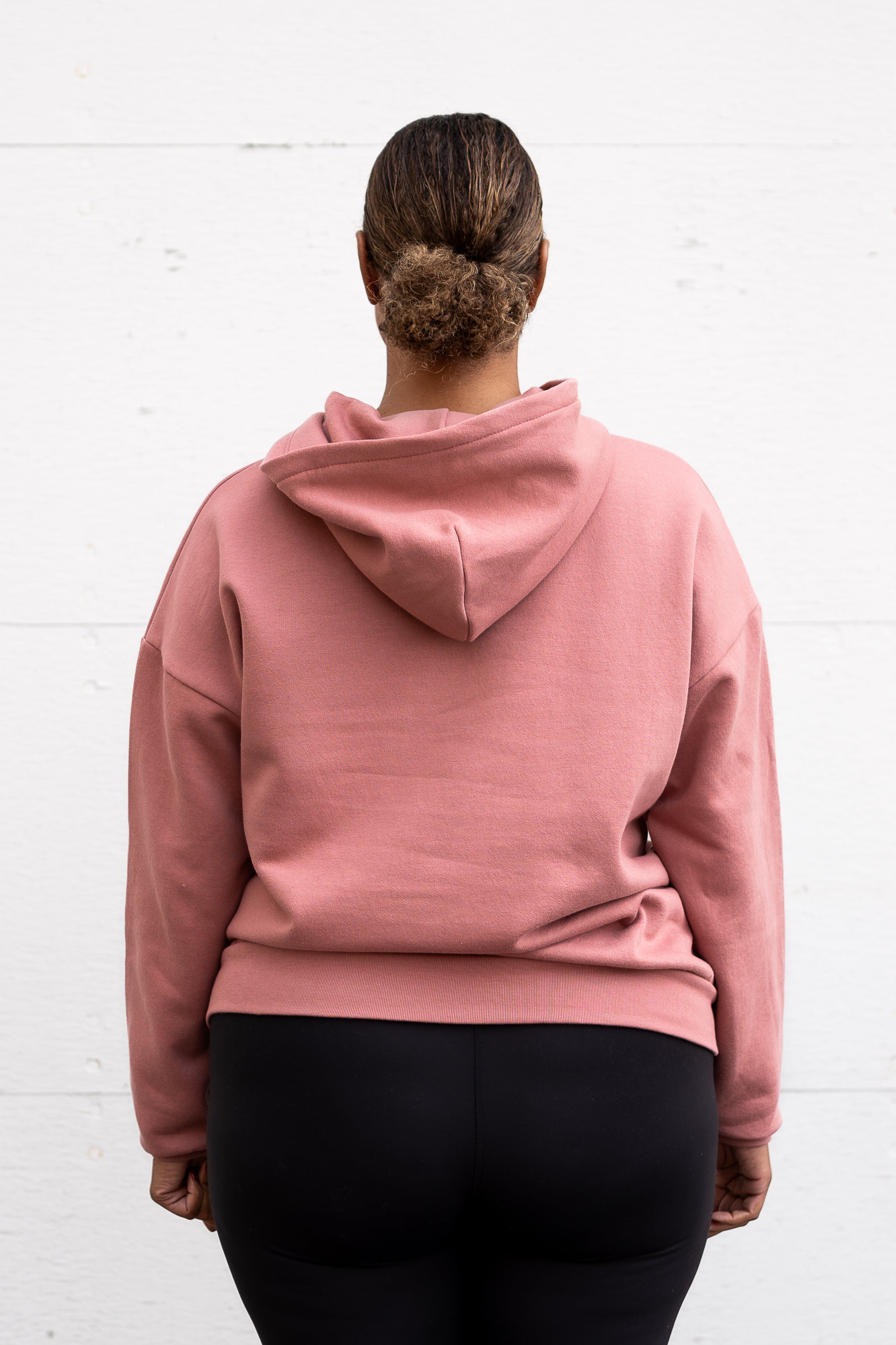 Woman standing facing backwards wearing a unisex oversize bamboo-organic cotton fleece hoodie in dusty rose, with dropped shoulders and a large kangaroo pocket.