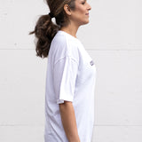 Woman standing facing sideways wearing a white unisex oversized t-shirt with a boxy fit  made from premium poly-cotton jersey, featuring the A$$piration Project logo on the front in grey, peach and green.