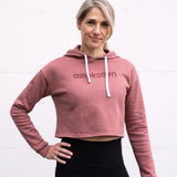 Woman standing facing forward with one hand on hip wearing a cropped bamboo-organic cotton fleece hoodie in dusty rose, featuring a finished hem, white drawstrings at the hood and the A$$piration Project logo on the front in tone on tone dark ruby pink.