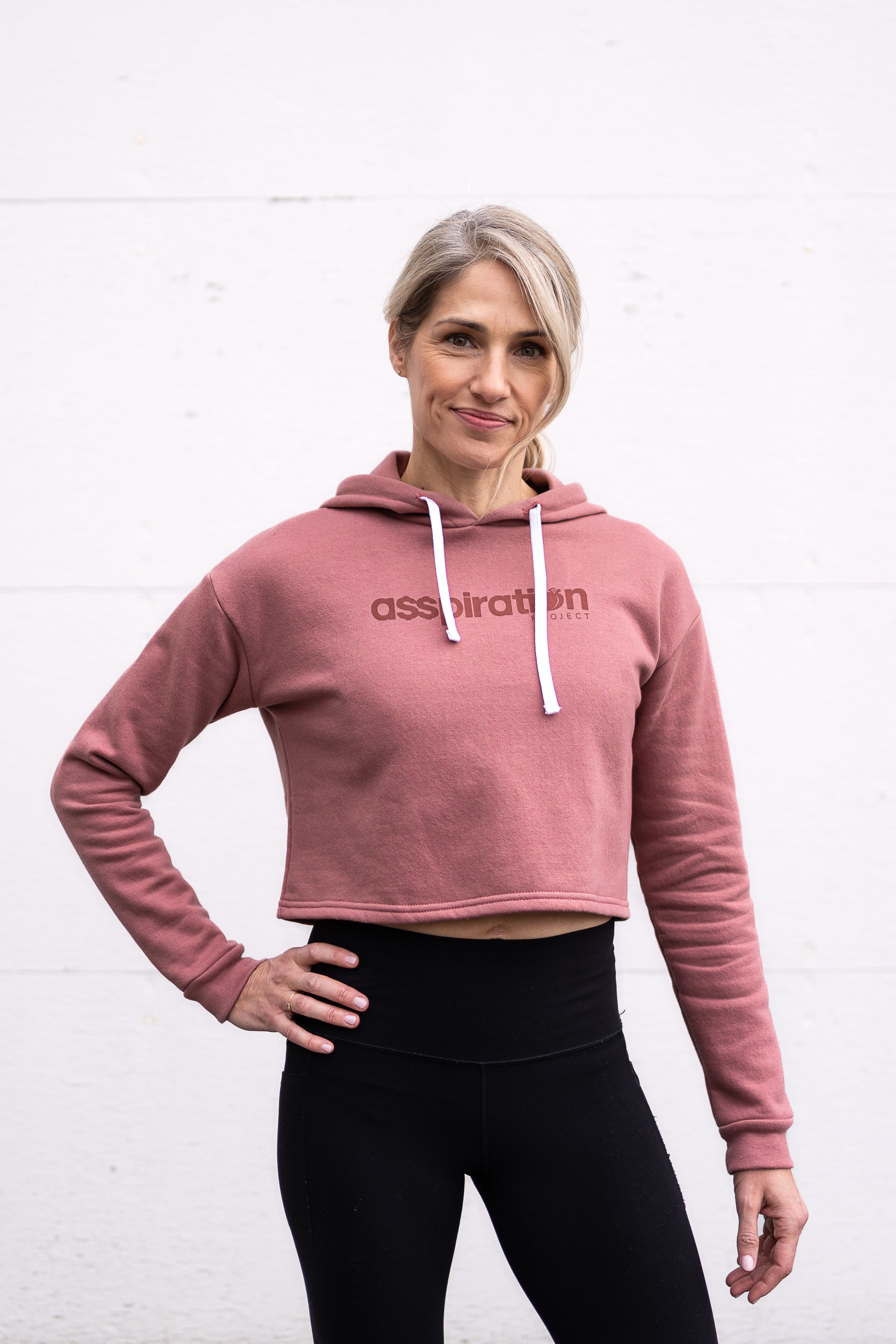 Woman standing facing forward with one hand on hip wearing a cropped bamboo-organic cotton fleece hoodie in dusty rose, featuring a finished hem, white drawstrings at the hood and the A$$piration Project logo on the front in tone on tone dark ruby pink.