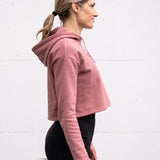 Woman standing facing sideways wearing a cropped bamboo-organic cotton fleece hoodie in dusty rose, featuring a finished hem, white drawstrings at the hood and the A$$piration Project logo on the front in tone on tone dark ruby pink.