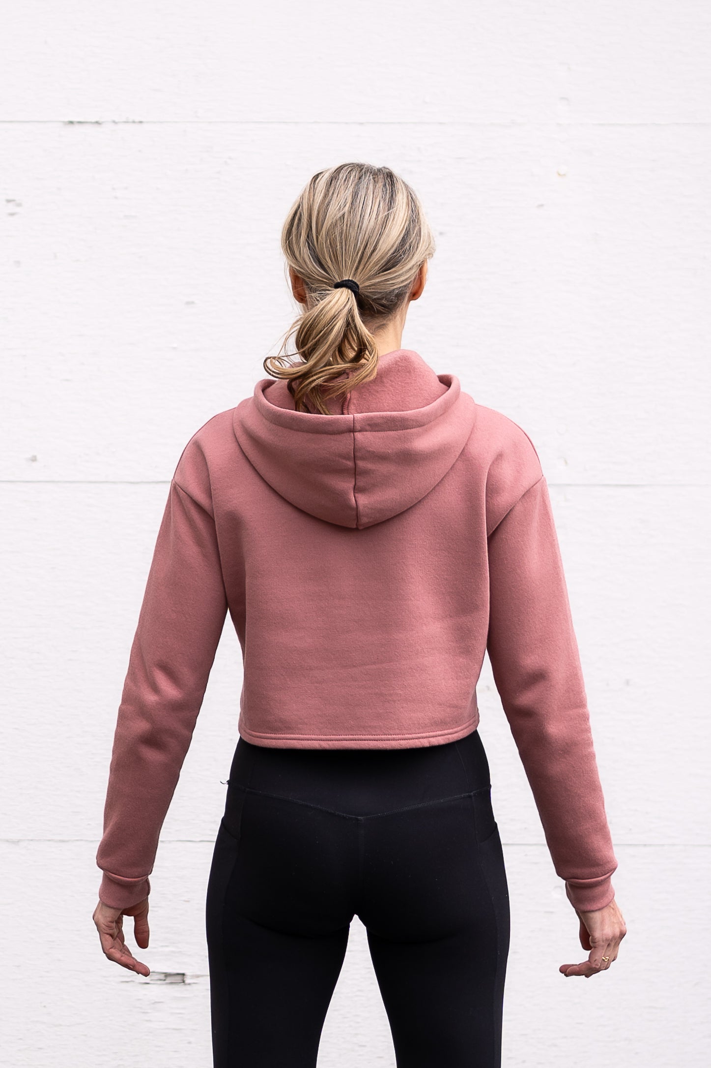 Woman standing facing backwards wearing a cropped bamboo-organic cotton fleece hoodie in dusty rose, featuring a finished hem.