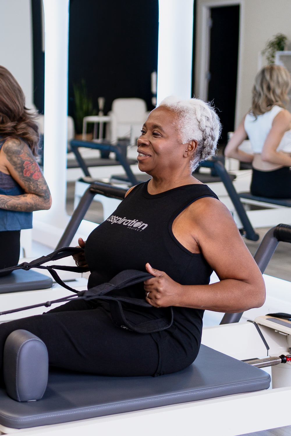 A woman seated upright on a pilates reformer machine, holding the straps in each hand in a rowing position, wearing a black full-length bamboo-organic cotton relaxed fit tank, featuring the A$$piration Project logo on the front in light grey.
