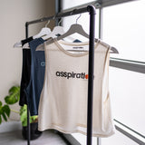 Three cropped bamboo-organic cotton tank tops in cream, slate blue and black, hanging from a bar in front of a garage window with a plant in the background. The front of the cream tank top features the A$$piration Project logo in grey, peach and green, the slate blue tank top features the A$$piration Project logo in light blue, and the black tank top features the the A$$piration Project logo in light grey.