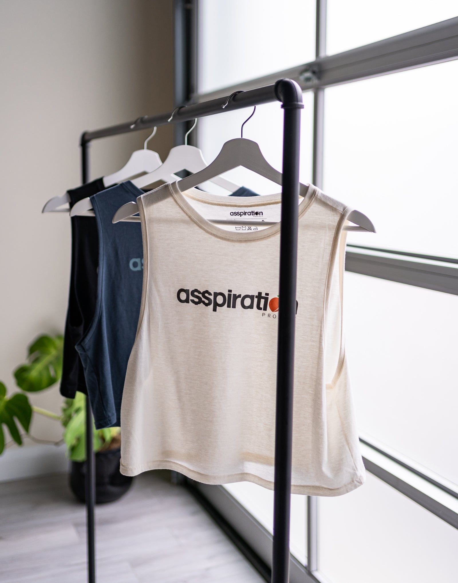 Three cropped bamboo-organic cotton tank tops in cream, slate blue and black, hanging from a bar in front of a garage window with a plant in the background. The front of the cream tank top features the A$$piration Project logo in grey, peach and green, the slate blue tank top features the A$$piration Project logo in light blue, and the black tank top features the the A$$piration Project logo in light grey.