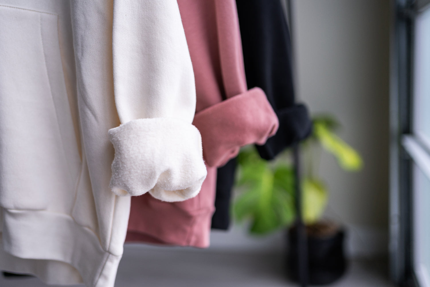 Close up of the rolled up sleeves of three unisex oversize bamboo-organic cotton fleece hoodies in cream, dusty rose and black, with a garage window and plant in the background.