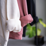 Close up of the rolled up sleeves of three unisex oversize bamboo-organic cotton fleece hoodies in cream, dusty rose and black, with a garage window and plant in the background.