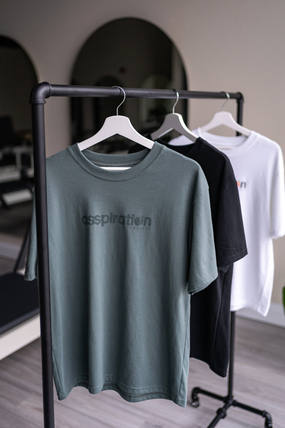 Three unisex oversized t-shirts  in sage green, black and white with a boxy fit made from premium poly-cotton jersey, featuring the A$$piration Project logo on the front, hanging from a bar in front of a mirror.