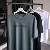 Three unisex oversized t-shirts  in sage green, black and white with a boxy fit made from premium poly-cotton jersey, featuring the A$$piration Project logo on the front, hanging from a bar in front of a mirror.