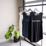 Three full length tanktops in black, slate blue and cream, with relaxed fit and rounded hems, hanging from a bar in front of a garage window with a plant in the background.