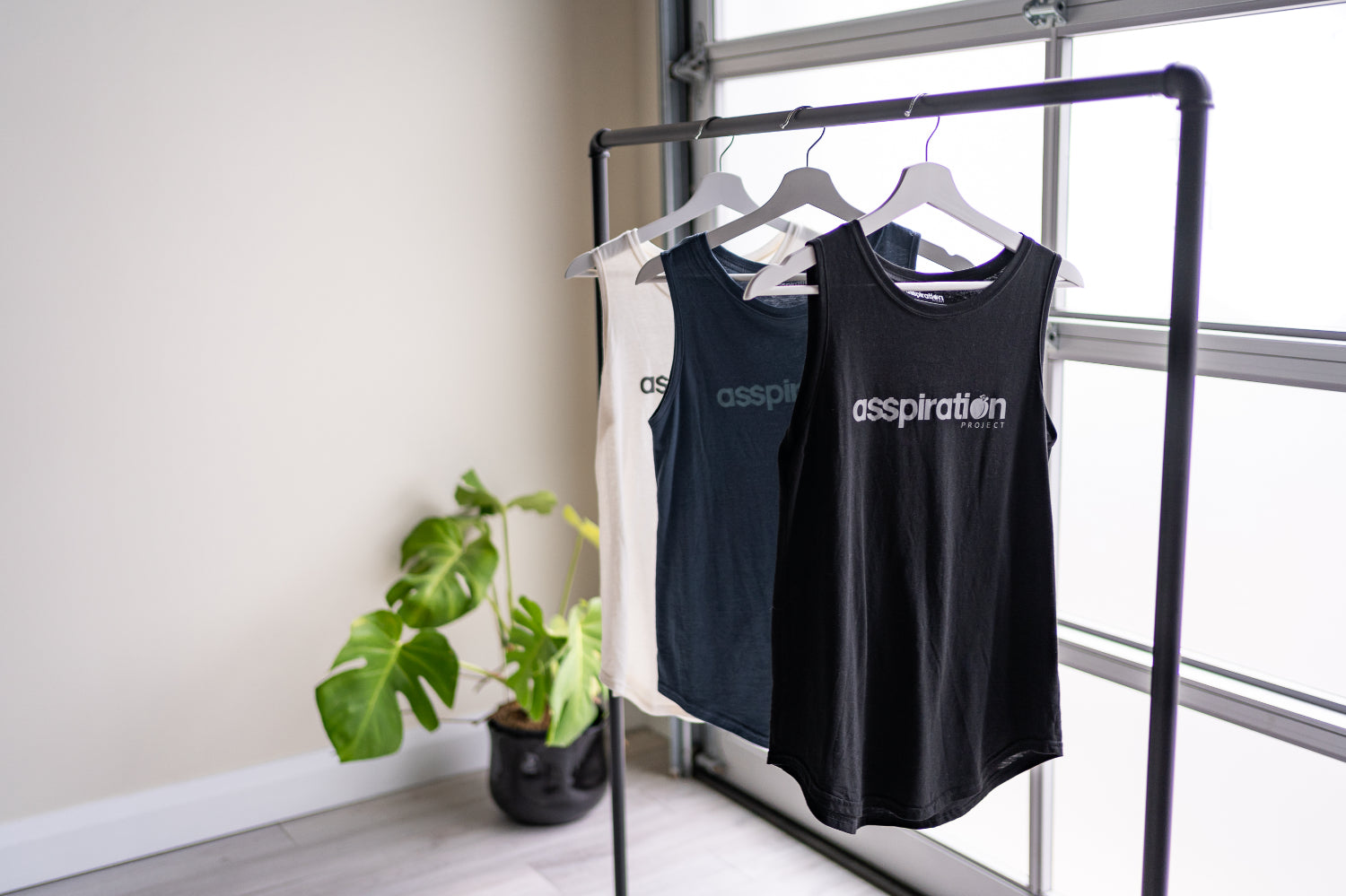 Three full length tanktops in black, slate blue and cream, with relaxed fit and rounded hems, hanging from a bar in front of a garage window with a plant in the background.