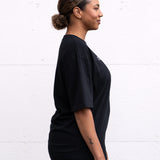Woman standing facing sideways wearing a black unisex oversized t-shirt with a boxy fit, made from premium poly-cotton jersey, featuring the A$$piration Project logo on the front in light grey.