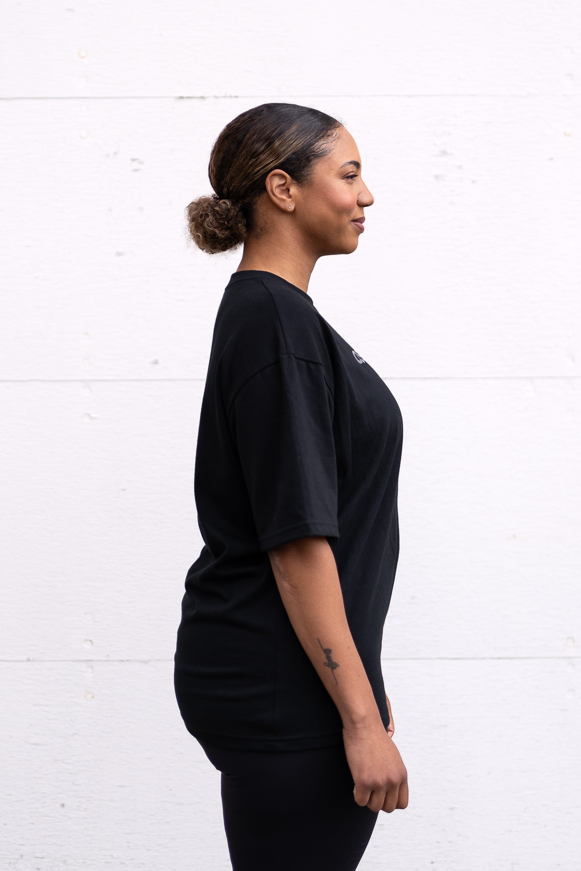 Woman standing facing sideways wearing a black unisex oversized t-shirt with a boxy fit, made from premium poly-cotton jersey, featuring the A$$piration Project logo on the front in light grey.