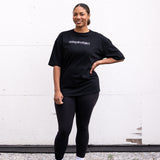 Woman standing facing forward with one hand on hip, wearing a black unisex oversized t-shirt with a boxy fit, made from premium poly-cotton jersey, featuring the A$$piration Project logo on the front in light grey.