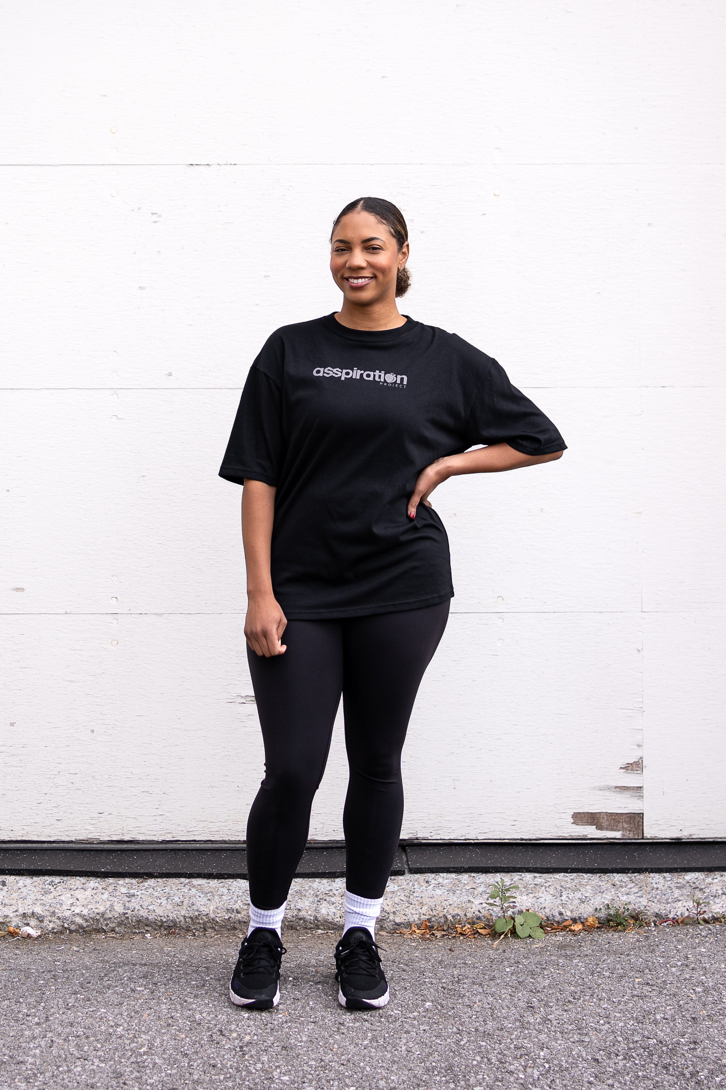 Woman standing facing forward with one hand on hip, wearing a black unisex oversized t-shirt with a boxy fit, made from premium poly-cotton jersey, featuring the A$$piration Project logo on the front in light grey.