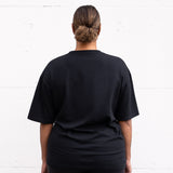 Woman standing facing backwards wearing a black unisex oversized t-shirt with a boxy fit, made from premium poly-cotton jersey.