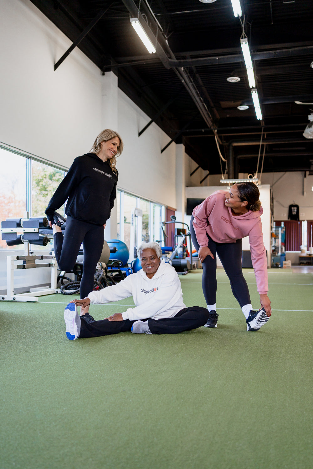 Three women stretching in a gym, wearing unisex oversize bamboo-organic cotton fleece hoodies in black, cream and dusty rose with dropped shoulders and a large kangaroo pocket, featuring the A$$piration Project logo on the front.