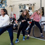 Three women in a gym engaged in "tug of war" with battle ropes, wearing cropped bamboo-organic cotton hoodies in cream, black and dusty rose, all featuring the A$$piration Project logo on the front.
